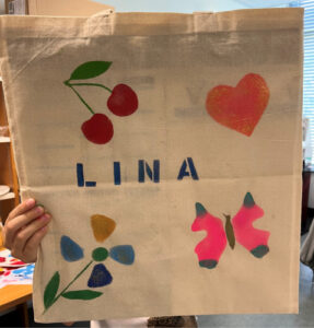 Des main tiennent un sac en tissu sur lequel est dessiné en pochoir le mot LINA, des cerises, un cœur, un papillon et une fleur