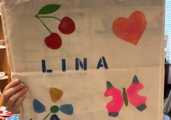 Des main tiennent un sac en tissu sur lequel est dessiné en pochoir le mot LINA, des cerises, un cœur, un papillon et une fleur